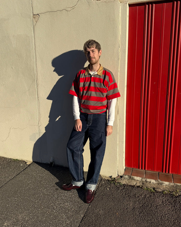 A man stands outdoors against a beige wall and a red corrugated metal door, wearing a red and gray striped short-sleeve polo shirt over a white long-sleeve crewneck, dark blue jeans, and burgundy loafers.