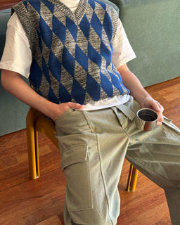 A person seated on a yellow chair wearing a blue and gray argyle knit sweater vest over a white T-shirt, with olive cargo pants, holding a metal cup in a home interior setting.