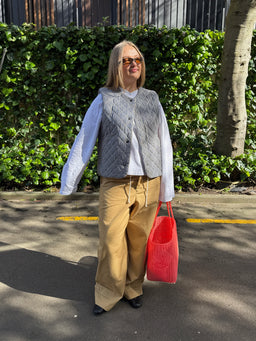 A woman stands outdoors on a paved path in front of a green hedge, wearing a gray quilted vest over a white long-sleeve shirt, tan wide-leg pants, and holding a bright pink woven tote bag.