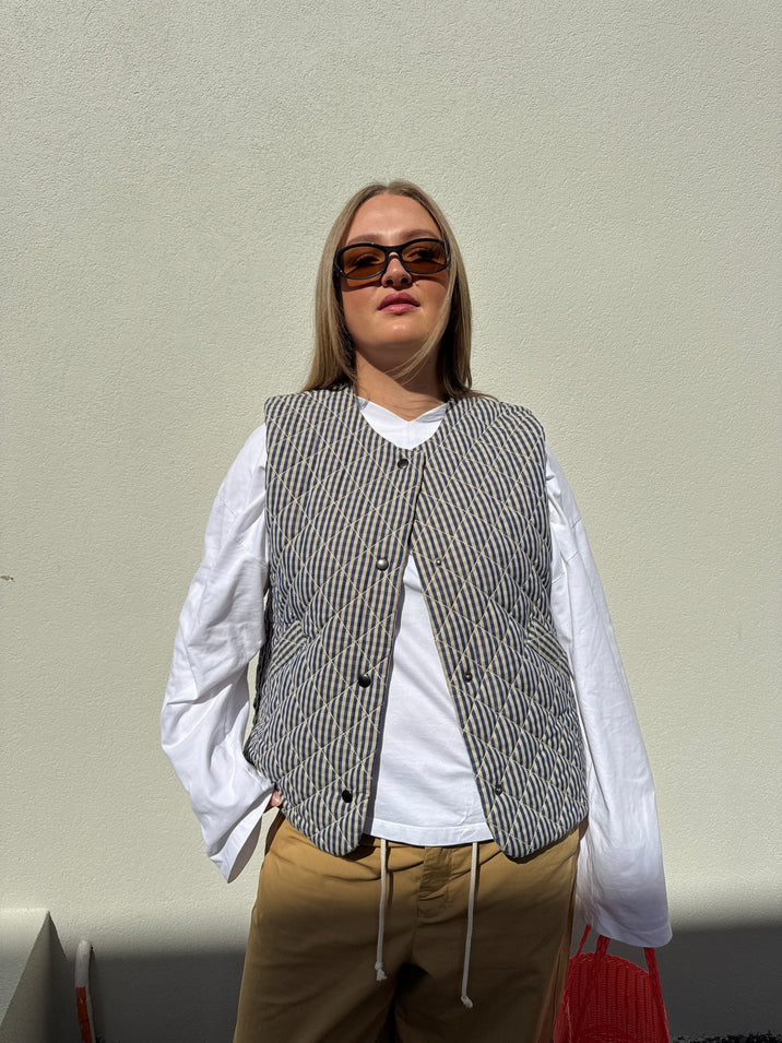 A woman wearing a gray and white striped quilted vest over a white long-sleeve shirt, tan pants, and dark sunglasses, standing against a plain light beige wall.