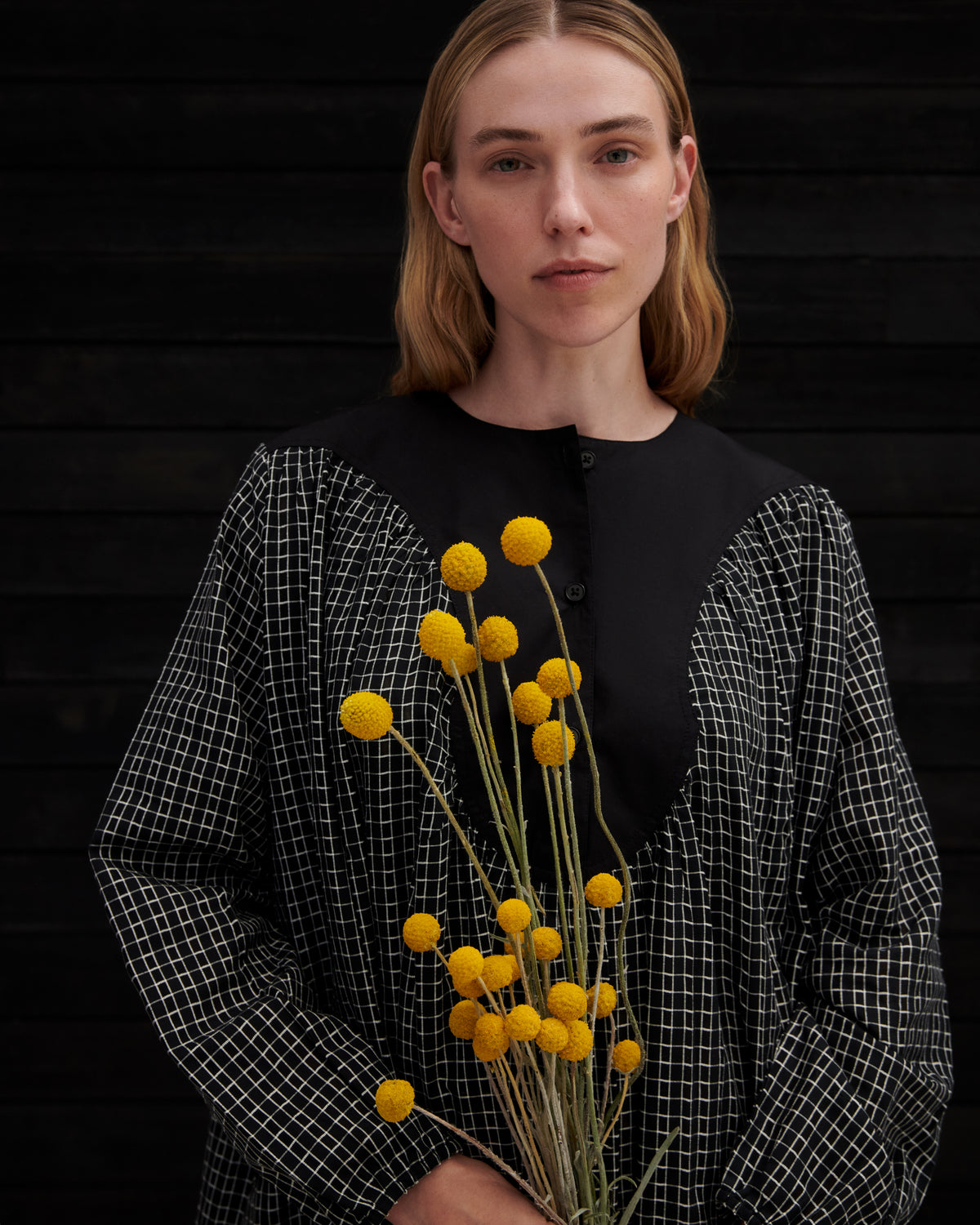 A woman wearing a black top with a black-and-white grid-patterned long-sleeve layer, holding a bunch of yellow globe flowers against a dark wood background.