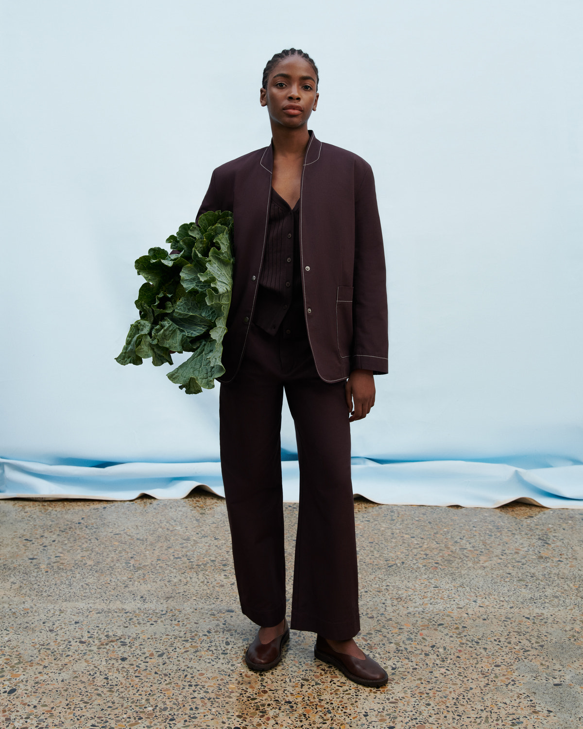 A model stands against a light blue backdrop wearing a dark brown tailored suit with a blazer, vest, and wide-leg trousers, holding a bunch of green leafy vegetables.