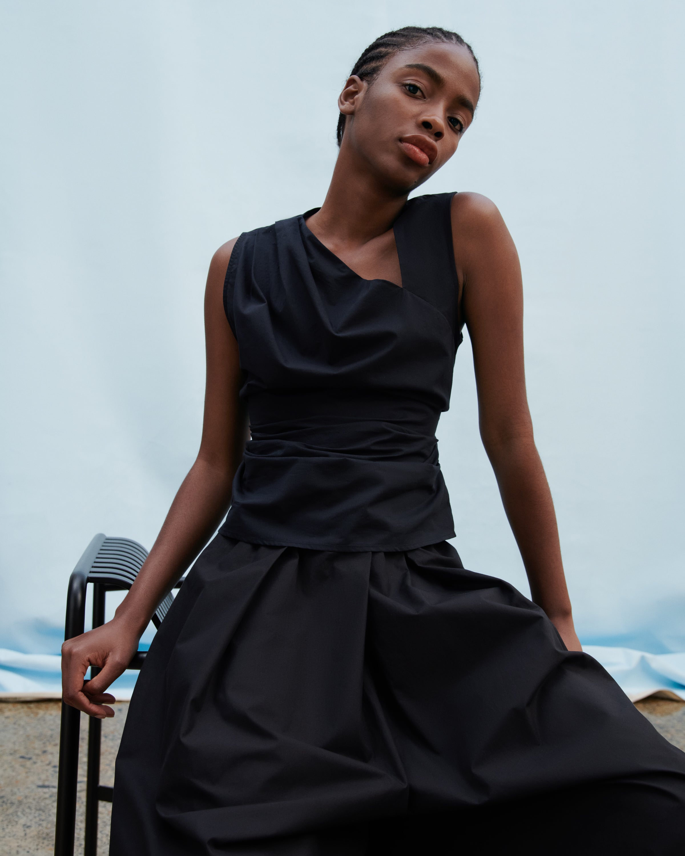A model wearing a black asymmetrical draped top and matching pleated skirt, seated on a black chair against a light blue backdrop.