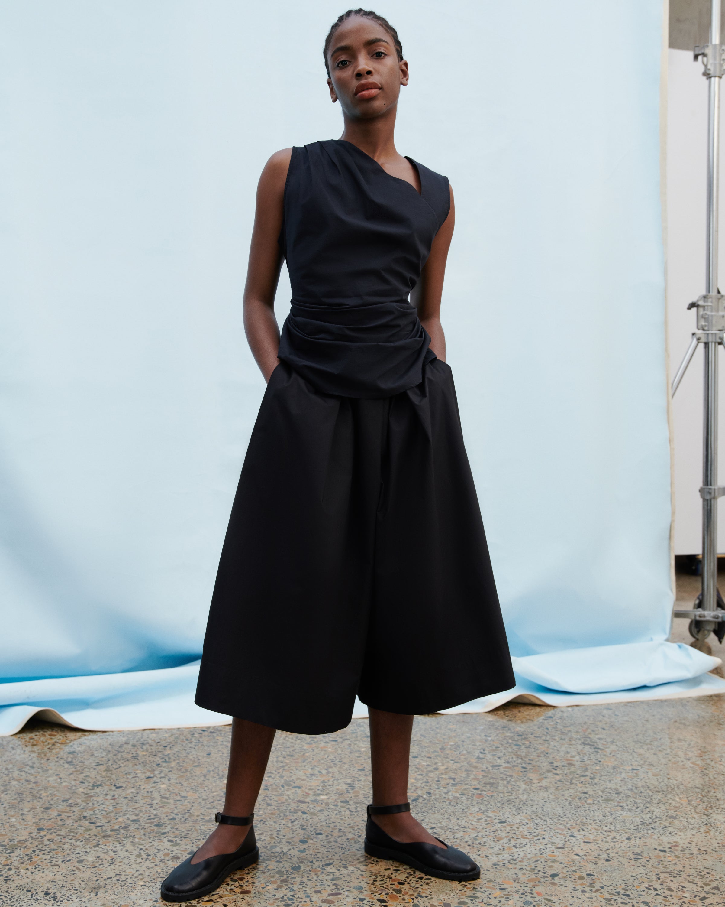 A model wearing a black sleeveless draped top and matching wide-leg cropped skirt, paired with black Mary Jane shoes, standing in front of a light blue studio backdrop.