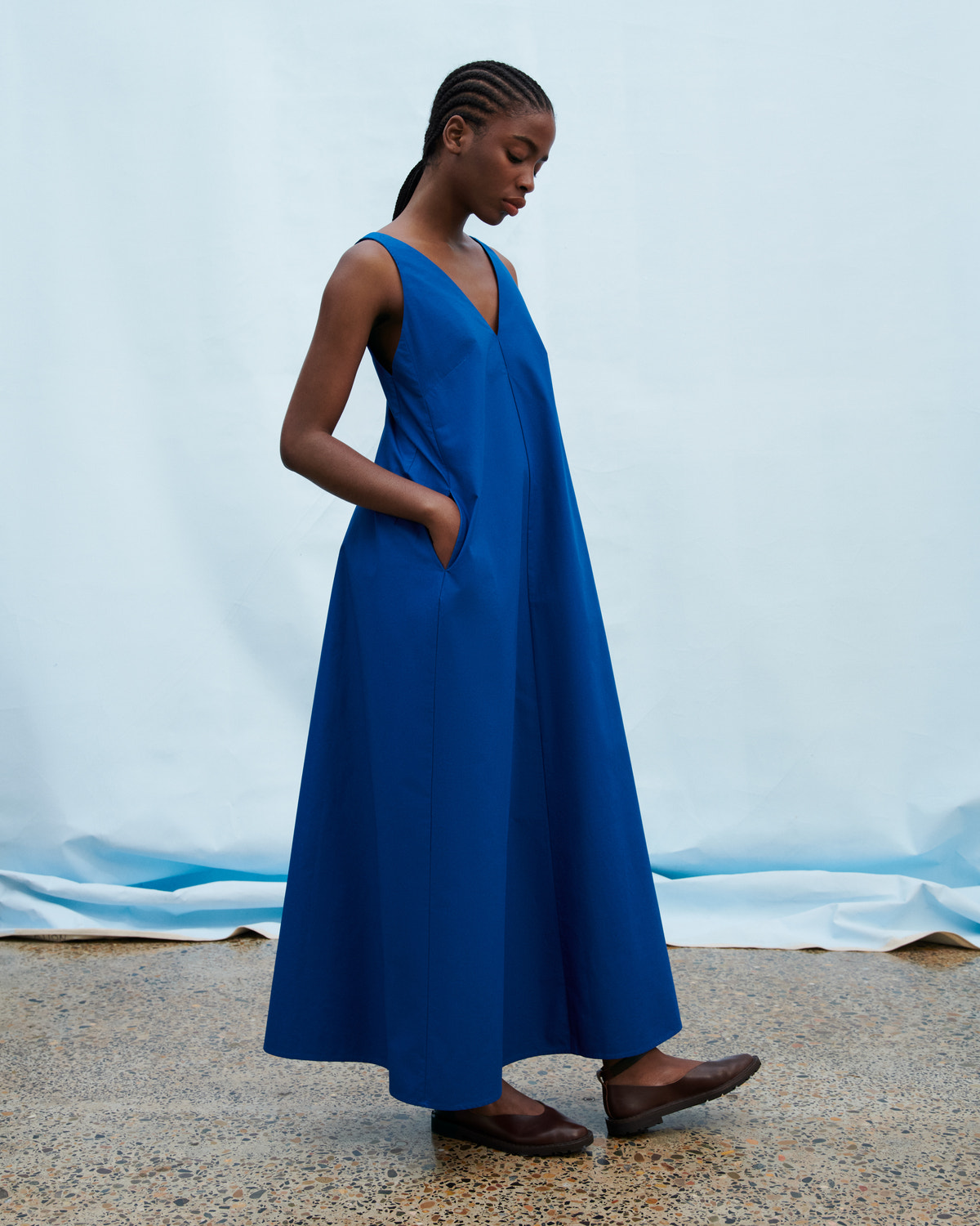 A woman wearing a sleeveless, V-neck, maxi-length blue dress with side pockets, standing against a light blue backdrop.