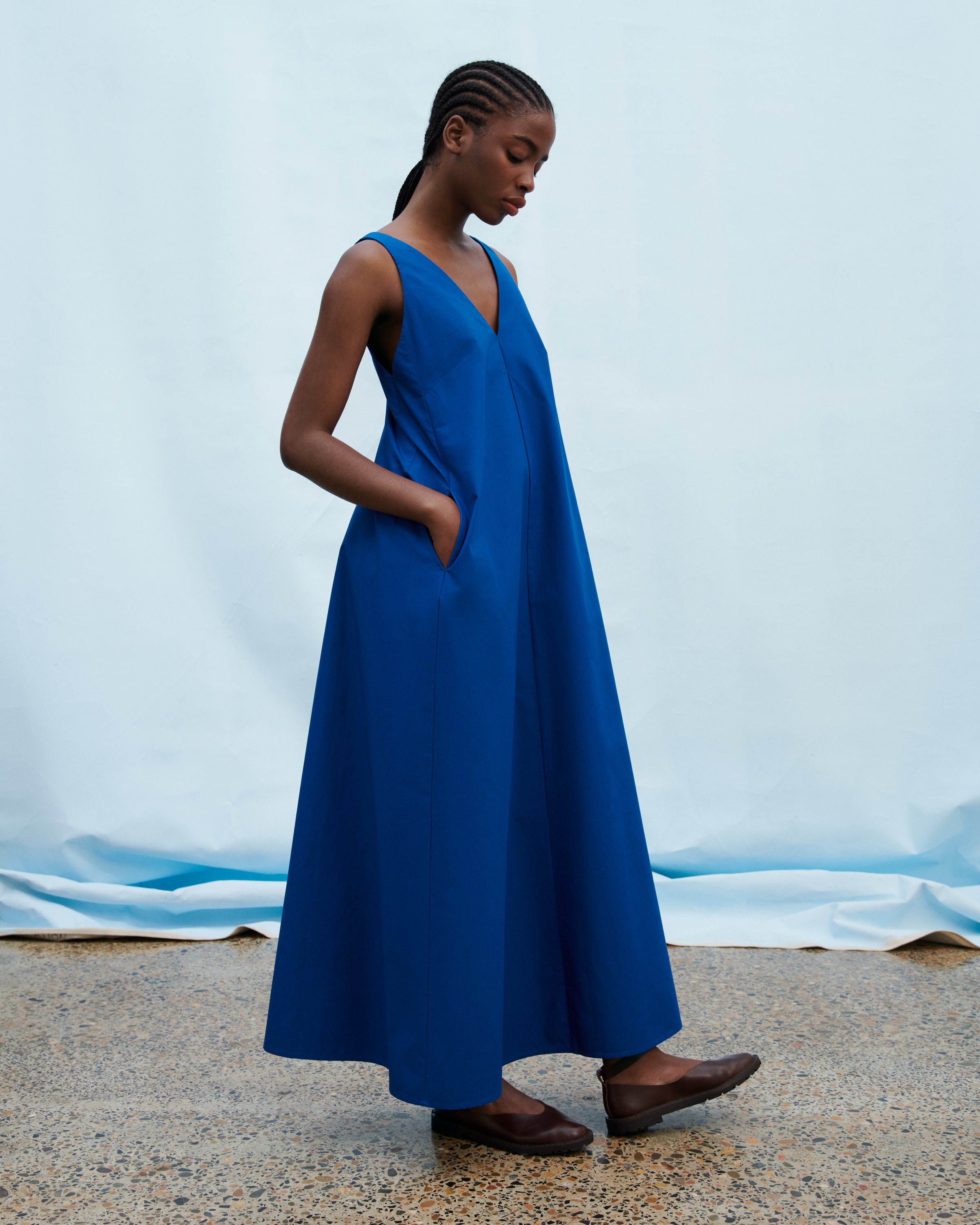 A woman wearing a sleeveless, V-neck, maxi-length blue dress with side pockets, standing against a light blue backdrop.
