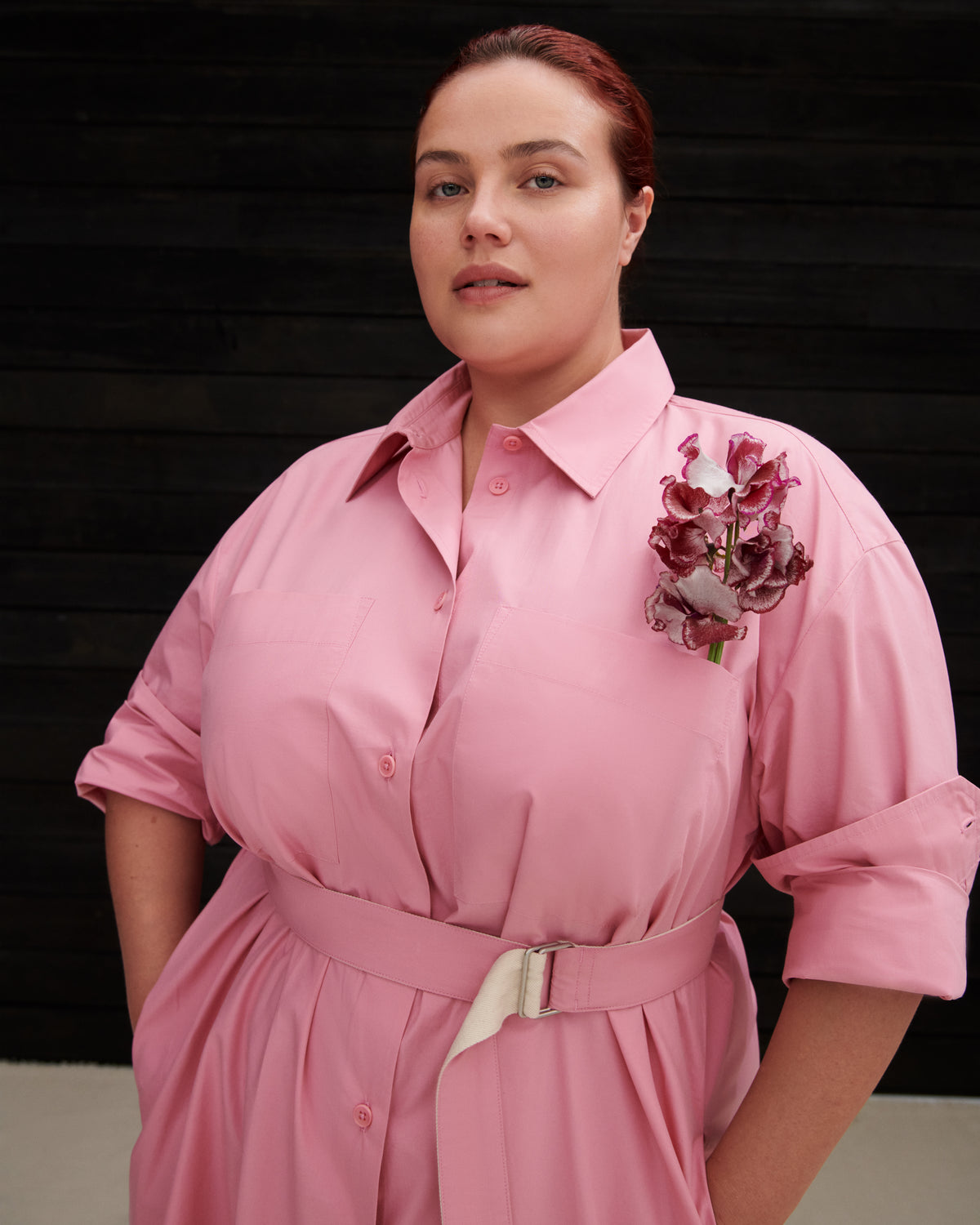 A woman wearing a pink collared shirt dress with rolled sleeves, a matching belt, and a burgundy floral brooch, posed against a dark wood-paneled background.