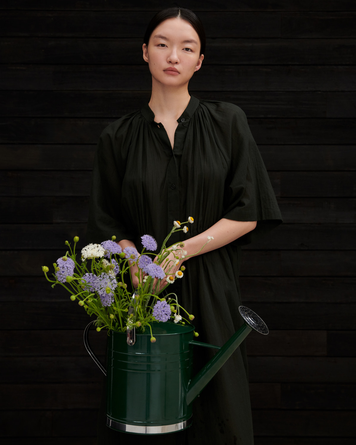 A woman wearing an olive green short-sleeve blouse stands against a dark wood plank wall, holding a green metal watering can filled with purple, white, and yellow wildflowers.