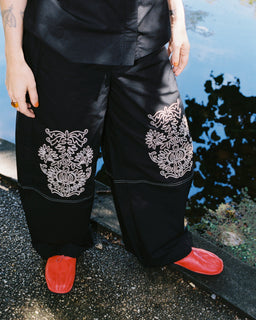 A person wearing black wide-leg pants with large white floral embroidery on the lower legs, paired with bright red shoes, standing outdoors near water.