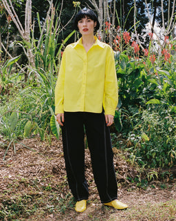 A person standing outdoors in a garden wearing a bright yellow button-up shirt, black wide-leg pants, and matching yellow shoes.