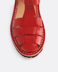 A close-up top-down view of a red leather sandal with a woven strap pattern and contrast stitching, set against a light gray background.