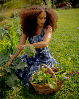 A woman with voluminous curly hair sits in a grassy garden, harvesting green broccoli from a plant while wearing a blue and white patterned dress. A wicker basket filled with leafy greens sits beside her.