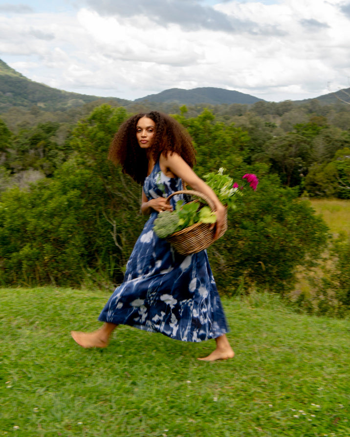 A woman with long curly hair walks barefoot across a grassy field, wearing a blue and white tie-dye maxi dress and carrying a wicker basket filled with green vegetables and pink flowers, with mountains and a cloudy sky in the background.