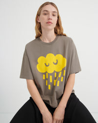 A woman wearing an oversized taupe-gray t-shirt with a yellow cartoon cloud and rain graphic, paired with black pants, seated against a light gray studio background.