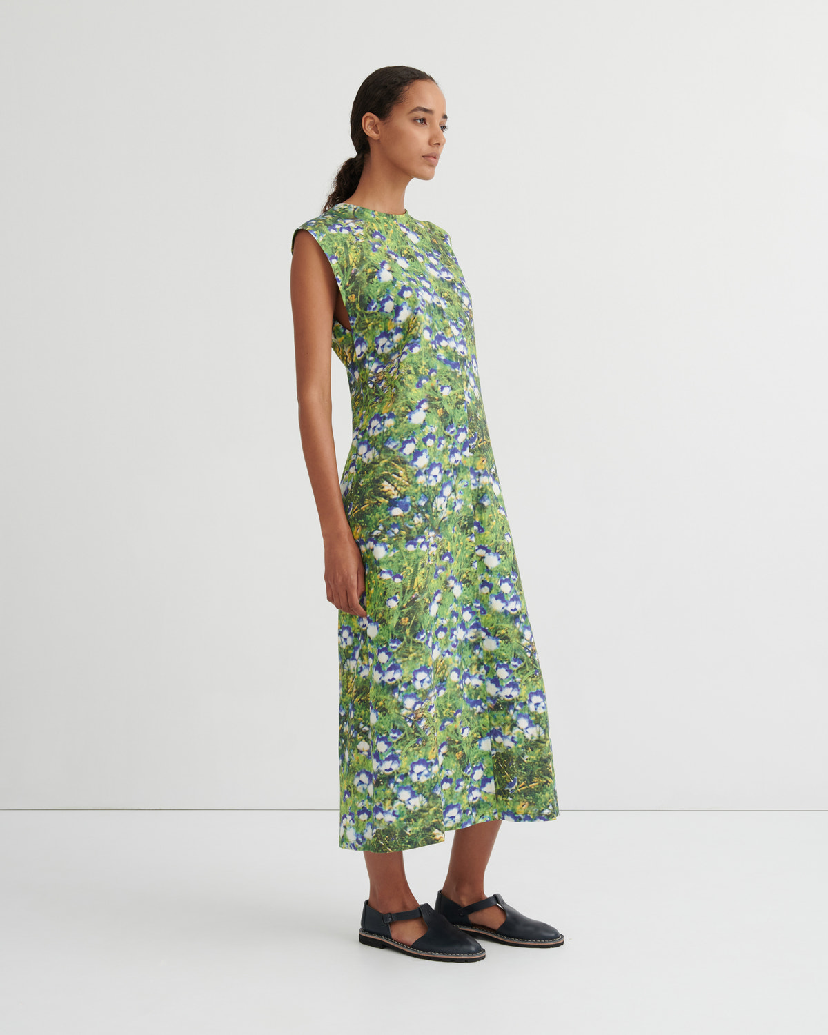 A woman modeling a sleeveless, knee-length green dress with a dense blue and white floral print, paired with black Mary Jane flats, against a plain white studio background.