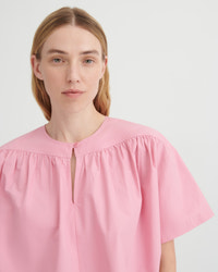 A woman wearing a light pink short-sleeve blouse with a gathered round neckline and a small front keyhole cutout, photographed against a plain light gray studio background.