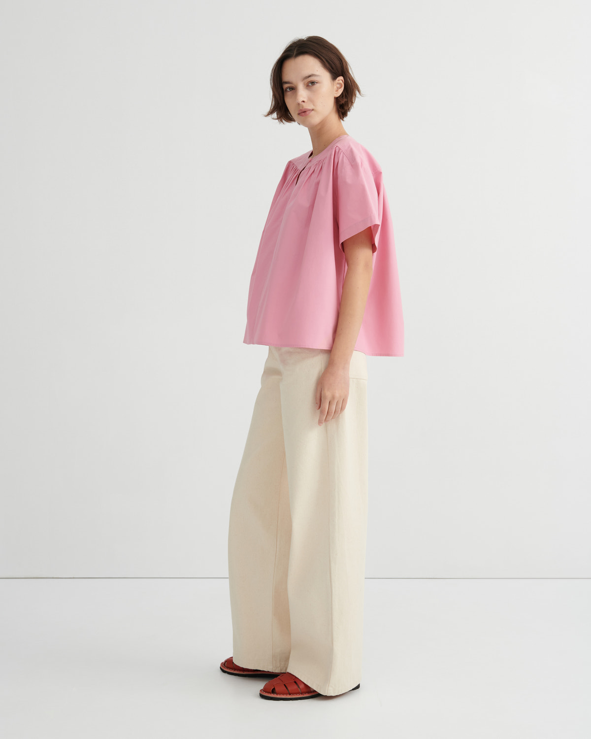 A model stands in profile wearing a light pink short-sleeve blouse with a gathered neckline, paired with wide-leg cream trousers and red sandals, against a clean white studio background.