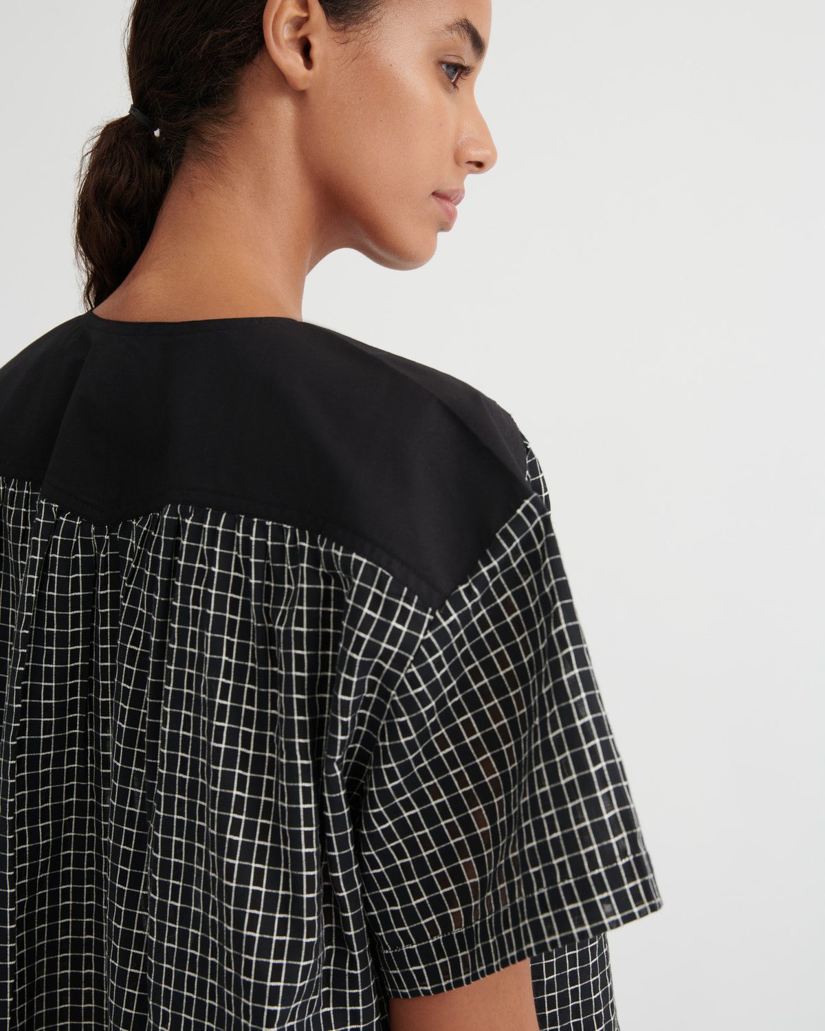 Close-up side profile of a woman wearing a loose black and white checkered top with a solid black yoke collar, against a plain light gray background.