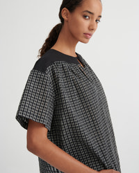 A woman wearing a black and white grid-patterned short-sleeve top with solid black shoulder yokes, photographed against a light gray studio background.
