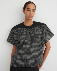 A woman modeling a short-sleeve top with a black and white grid pattern on the body and solid black shoulders and neckline, set against a plain light gray background.