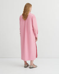 A woman viewed from behind wearing a long pink shirt dress with a collar and side slit, paired with beige sandals, against a plain white studio background.
