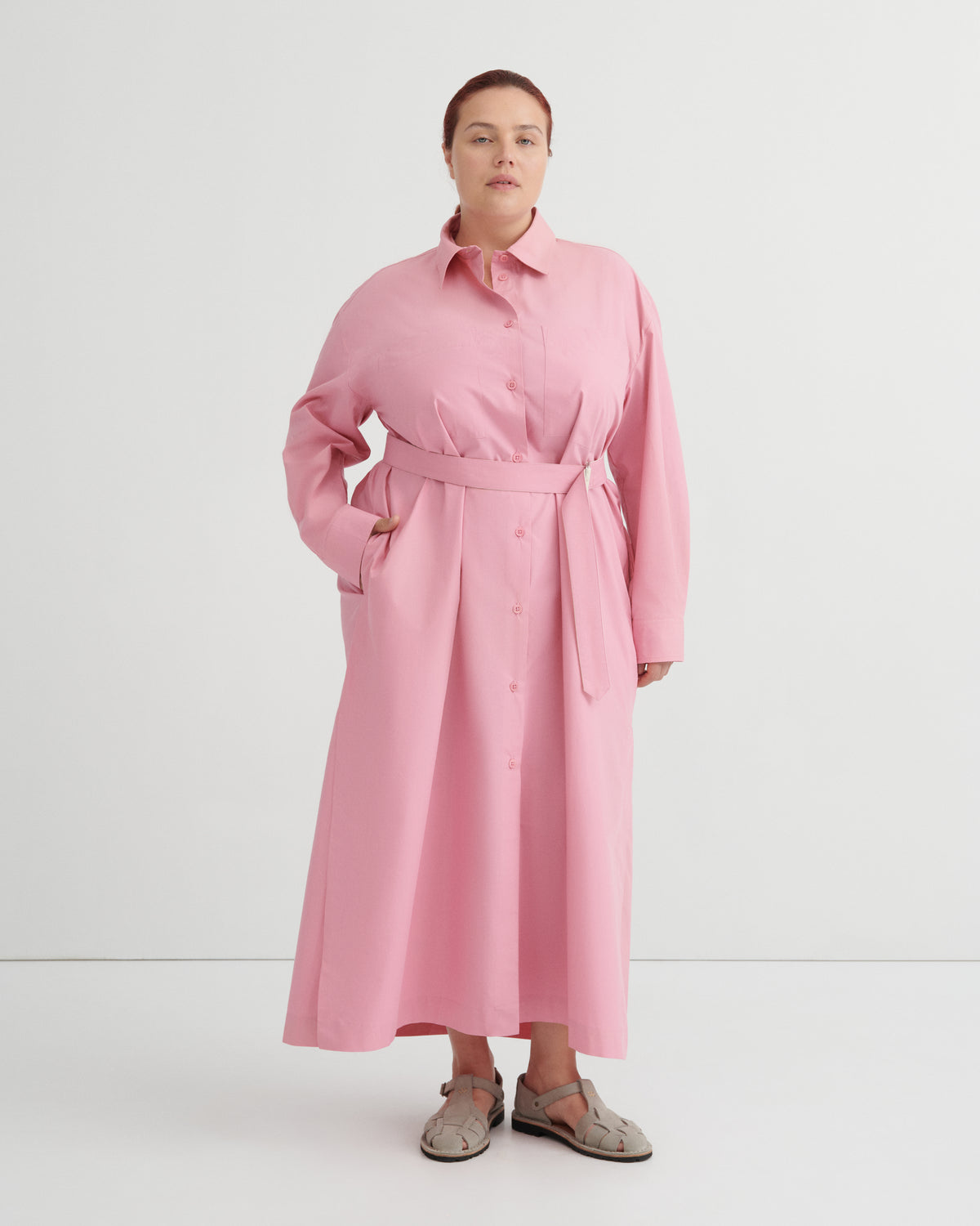 A woman modeling a pink long-sleeve shirt dress with a matching waist tie belt, standing against a plain white studio background.