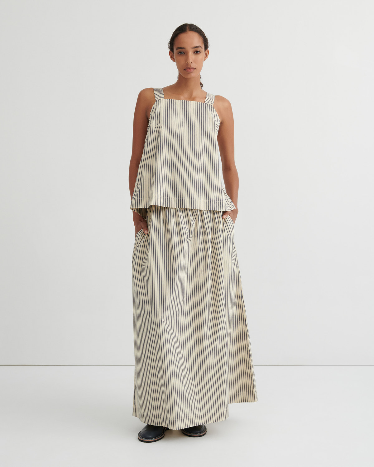 A model stands against a plain white studio background wearing a sleeveless beige and white vertically striped two-piece set consisting of a cropped top and wide-leg skirt.