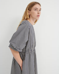 A woman wearing a loose-fitting, short-sleeved dress with vertical gray and white stripes, shown from a side profile against a plain light gray background.