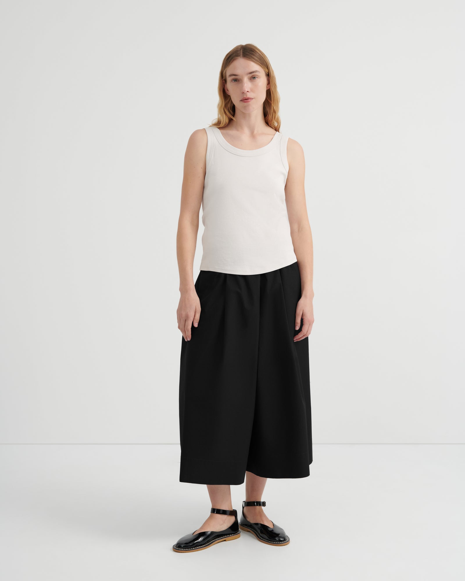 A woman wearing a sleeveless white ribbed tank top, loose black wide-leg cropped pants, and black flat sandals.