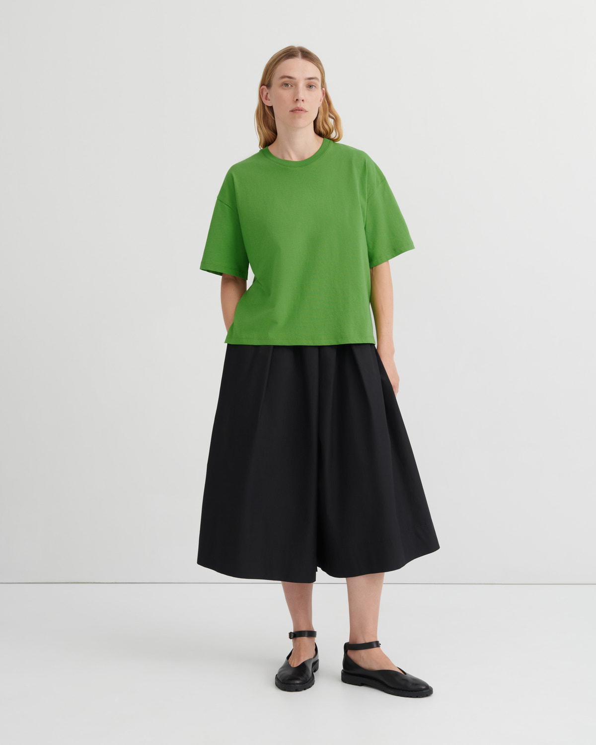 A model stands against a light gray studio background wearing a bright green short-sleeve crewneck top, a black pleated midi skirt, and black flat shoes.