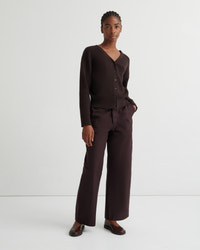 A model stands against a plain light gray studio background wearing a dark brown ribbed knit cardigan with a V-neck and buttons, matching wide-leg trousers, and dark brown loafers.