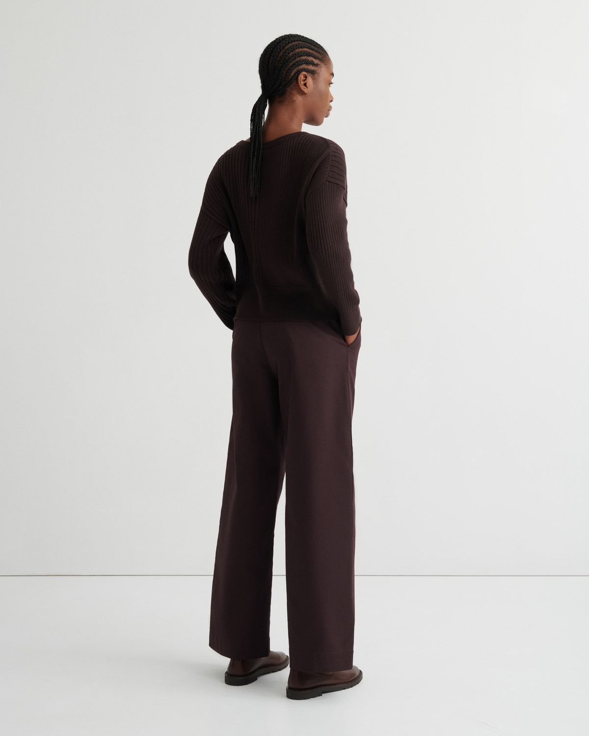 A model is shown from the back wearing a dark brown ribbed knit long-sleeve top and matching wide-leg trousers, standing against a clean white studio background.