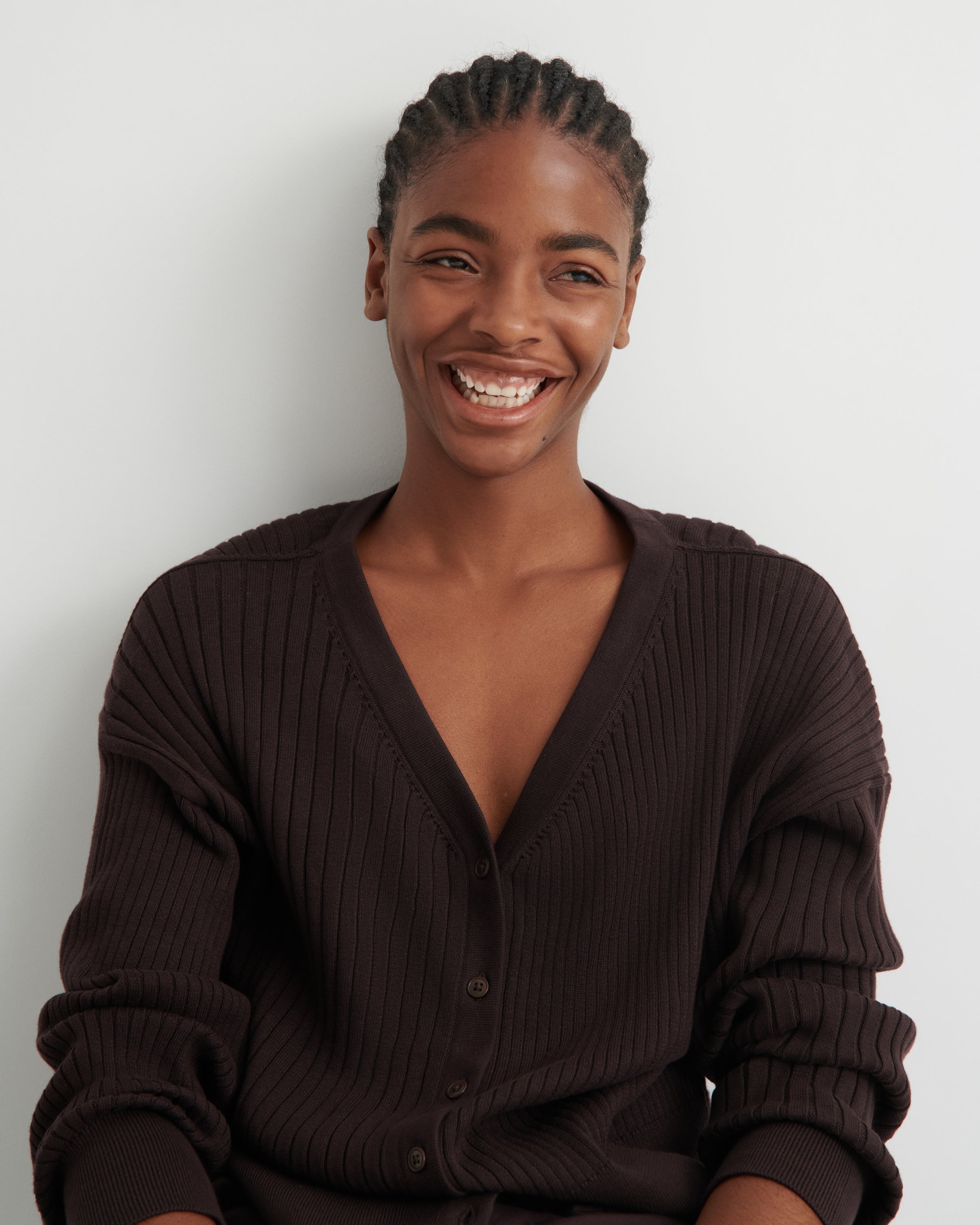 A smiling woman with braided hair wearing a dark brown ribbed cardigan sweater, photographed against a plain light gray background.