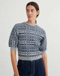 A woman wearing a short-sleeve knit top with a navy and white geometric grid pattern, paired with dark blue jeans, against a plain light gray studio background.