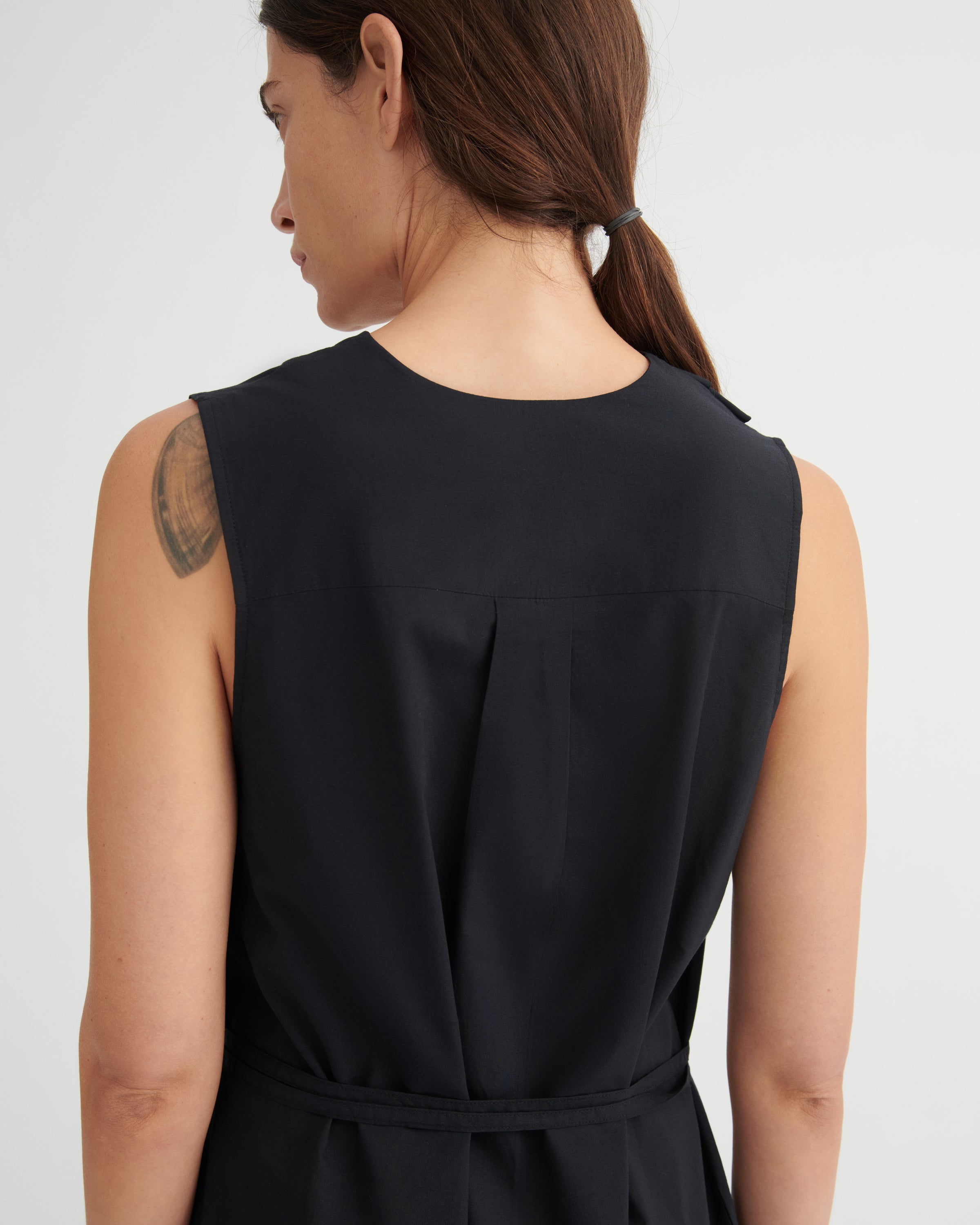 Rear view of a woman wearing a sleeveless black dress with a tie waist, shown against a plain light gray studio background.