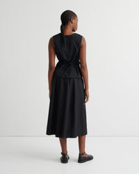A model viewed from the back wearing a sleeveless black midi dress with a tie waist, paired with black flat shoes, against a plain white studio background.