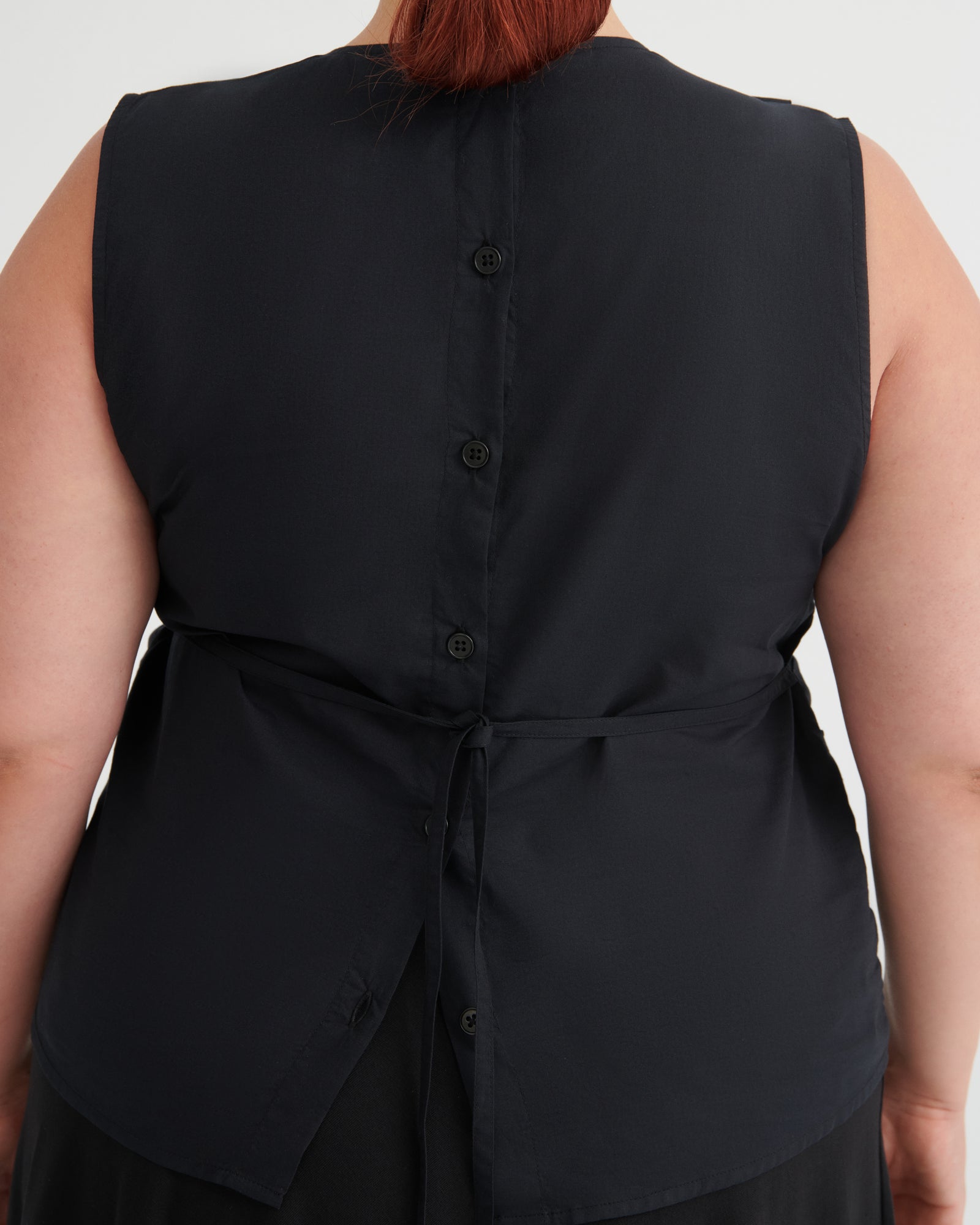 Close-up back view of a sleeveless black top with a buttoned back and tie detail, worn by a model against a light gray background.