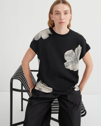 Person wearing a black t-shirt with floral designs sitting on a chair against a white background