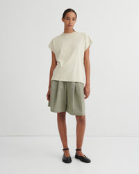 A model stands against a plain light gray studio background wearing a cream-colored short-sleeve t-shirt and olive green linen shorts, paired with black flat shoes.