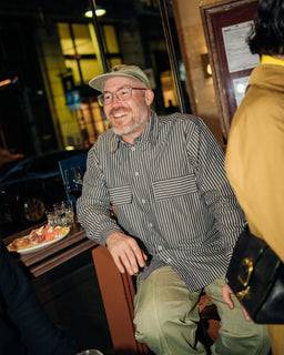 A man wearing a green baseball cap, glasses, a dark and light vertically striped button-down shirt, and olive green pants is smiling while sitting in a restaurant setting.