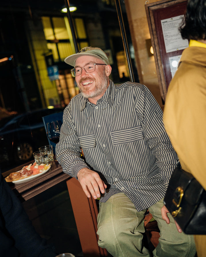 A man wearing a green baseball cap, glasses, a dark and light vertically striped button-down shirt, and olive green pants is smiling while sitting in a restaurant setting.