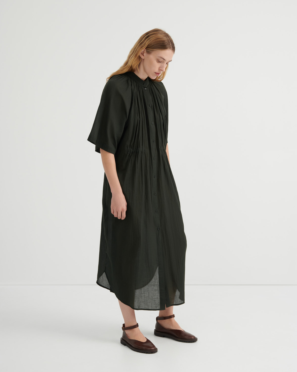 A woman modeling a loose, olive green midi dress with short flutter sleeves, a gathered high neckline, and a draped silhouette, paired with dark brown flat shoes against a plain white studio background.