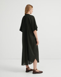 A woman stands in profile wearing a loose, flowing dark olive green dress with short wide sleeves and a draped silhouette, paired with brown flat shoes, against a clean light gray studio background.
