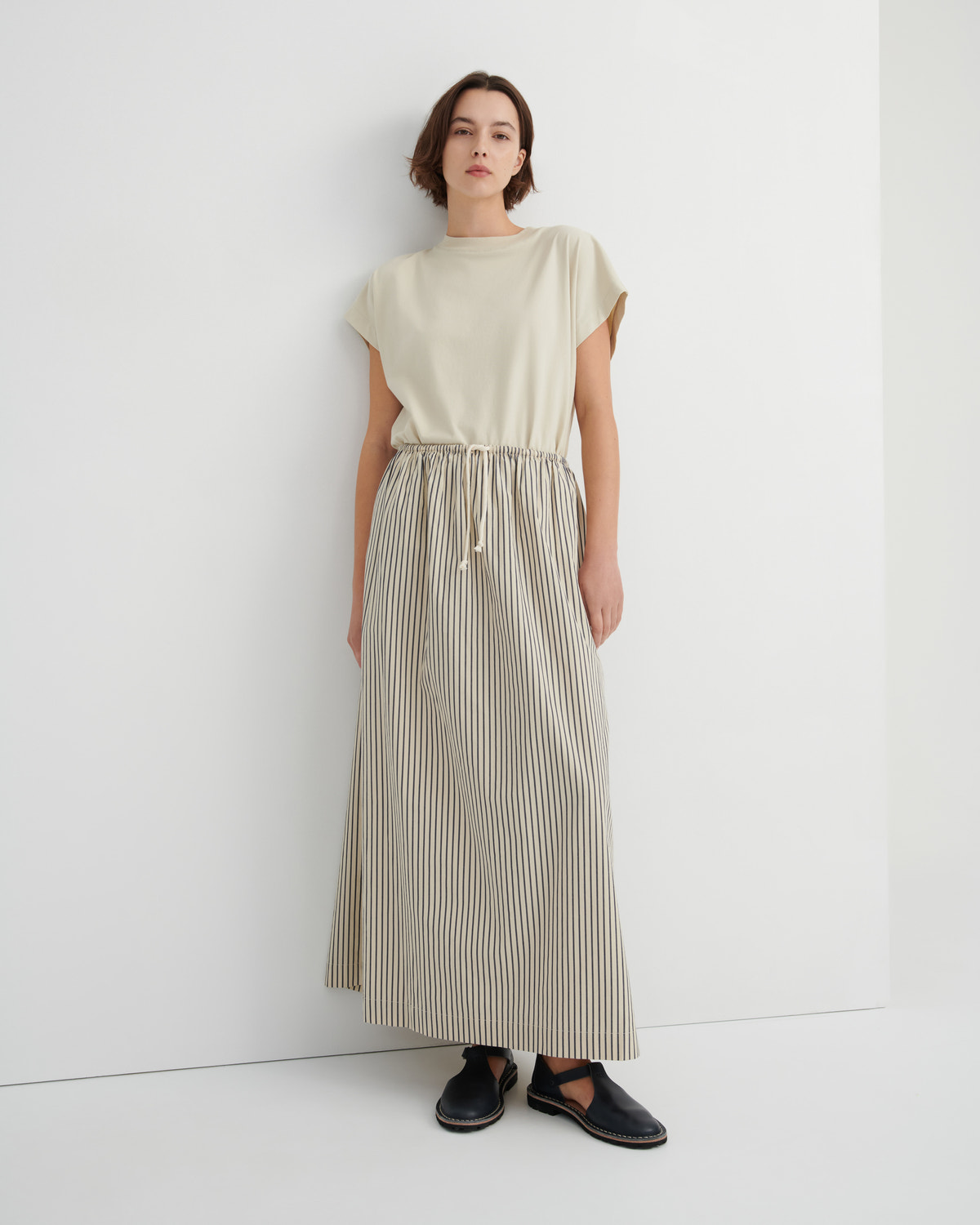 A model stands against a white wall wearing a cream-colored short-sleeve top and a matching beige and white vertically striped pleated maxi skirt with a drawstring waist, paired with black slip-on shoes.