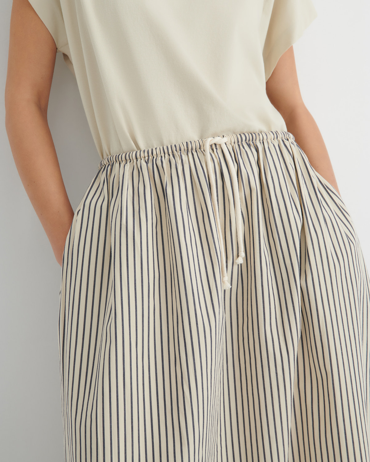 Close-up of a person wearing loose, wide-leg striped trousers with an elastic drawstring waist and a light cream top against a plain light gray background.