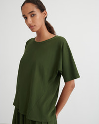 A woman wearing an olive green short-sleeve top with a relaxed, oversized fit, photographed against a plain light gray studio background.