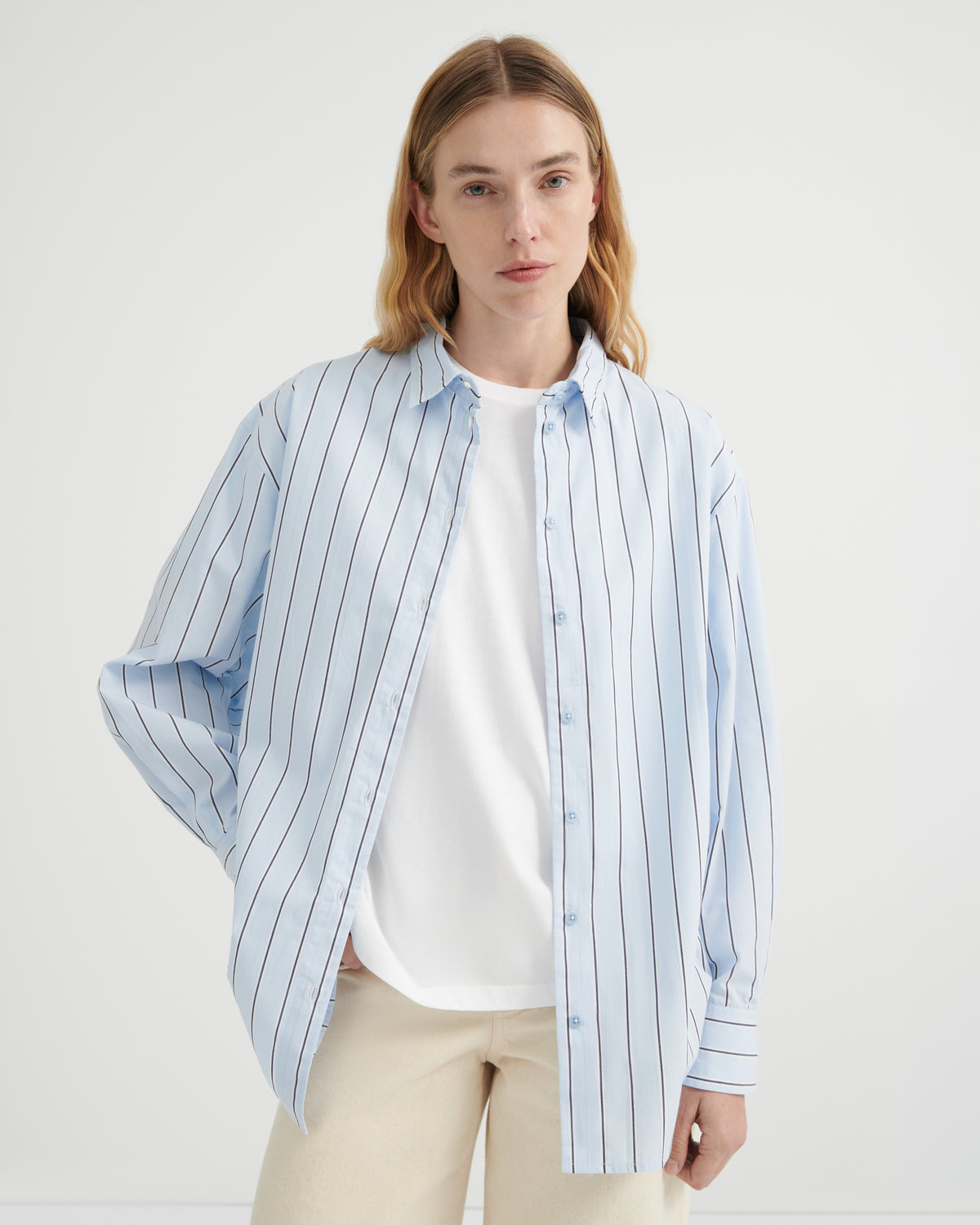 A woman wearing a light blue and white vertically striped button-up shirt over a white t-shirt and beige trousers.