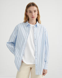A woman wearing a light blue and white vertically striped button-up shirt over a white t-shirt and beige trousers.