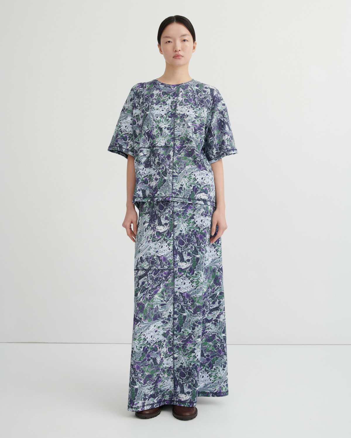 A model stands against a plain light gray studio background wearing a tee and maxi skirt with a blue, purple, and green abstract marbled pattern.