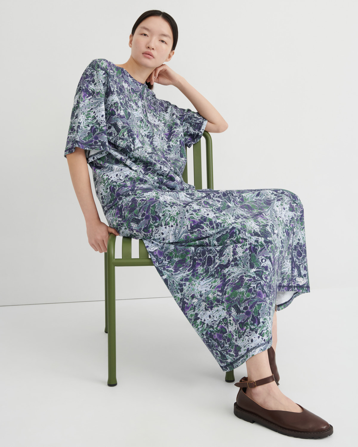 A model sits on a green metal chair wearing a loose, short-sleeved tee and maxi skirt with a blue, purple, and green abstract marbled print, paired with dark brown flat shoes, against a plain white studio background.